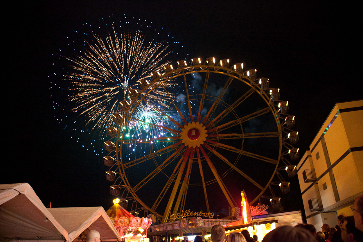 Ein Riesenrad, dahinter eine Feuerwerksblume am dunklen Nachthimmel.