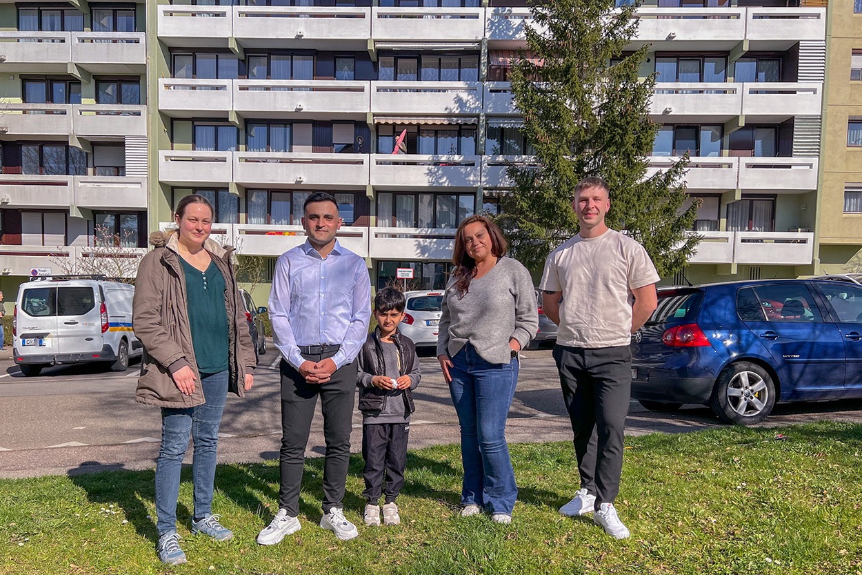 Eine Gruppe von Leuten steht vor einem Wohnblock und blickt in eine Kamera.