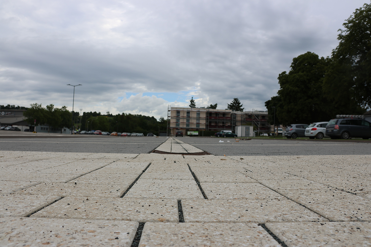 Pflastersteine, dahinter eine aspahltierrte Fläche, weiter hinten Gebäude