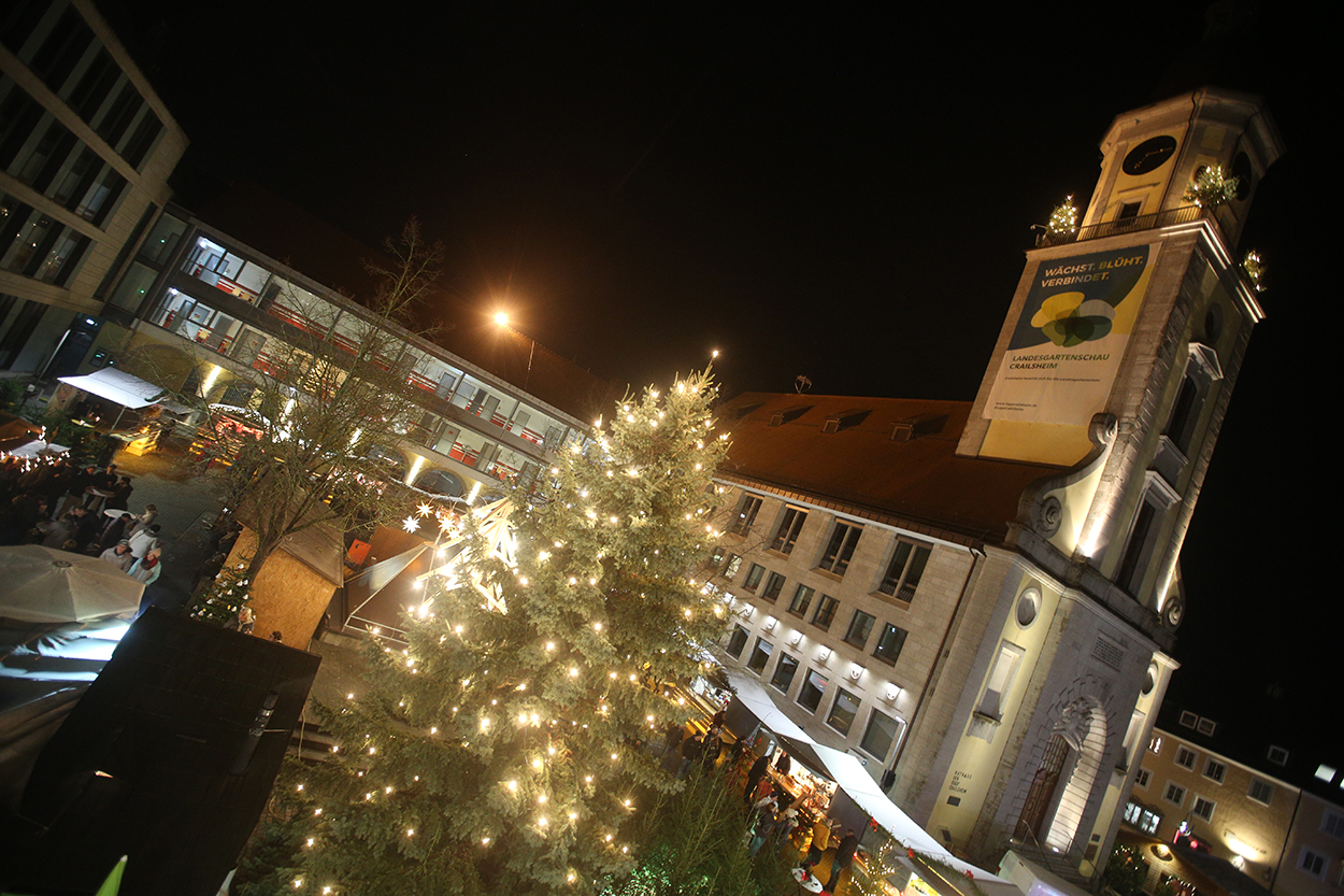 ein Foto in Schräglage mit leuchtendem Weihnachtsbaum vor einem Rathausturm mit Buden drumherum