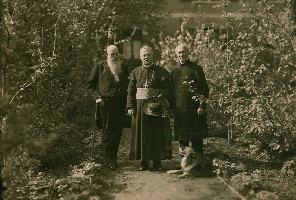 Drei Männer, würdig gekleidet, stehen in einem Garten und schauen in die Kamera